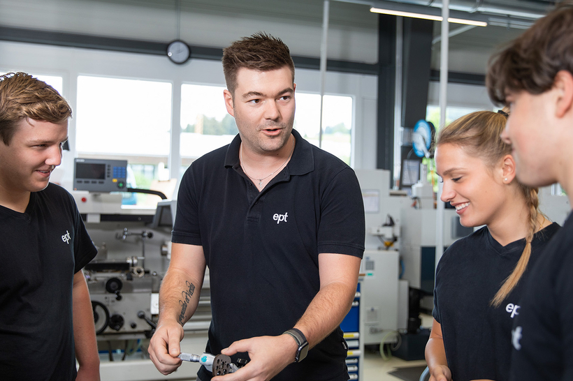 Auf dem Bild sind vier Personen in einem technischen Umfeld zu sehen, wobei ein Mann etwas erklärt und die anderen Teilnehmer aufmerksam zuhören.