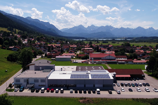 Auf dem Bild ist eine ländliche Szene zu sehen, die ein modernes Gebäude mit Photovoltaikanlagen sowie ein Wohngebiet vor einer Berglandschaft zeigt.